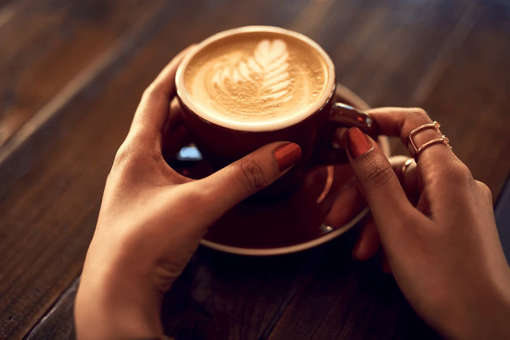 Hands holding a warm cup of latte with leaf-shaped latte art, resting on a wooden table.