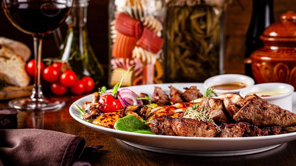 Alt text: A lavish plate of grilled meats and roasted vegetables served on a white dish, accompanied by small bowls of sauce. Fresh cherry tomatoes, bread, jars of pasta and pickled vegetables, and a glass of red wine sit in the background, creating a warm, rustic dining atmosphere.