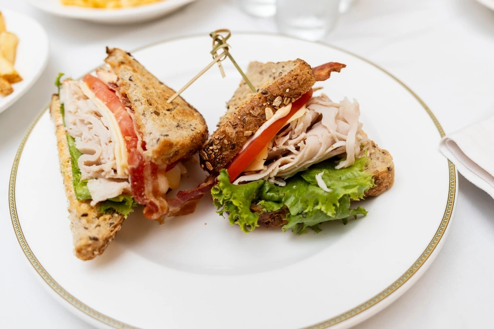 A gourmet sandwich served on multigrain bread, layered with sliced turkey, crispy bacon, fresh lettuce, tomato, and cheese, presented neatly on a white plate with a gold rim.